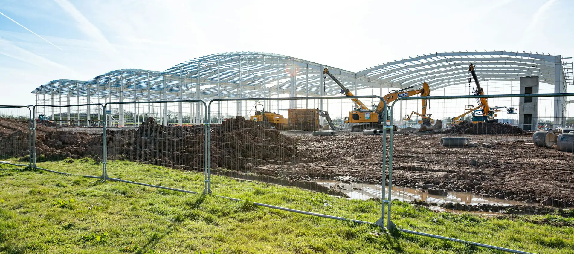Steel portal frames and roof purlins spanning a warehouse construction site, with cranes and earthmoving equipment.